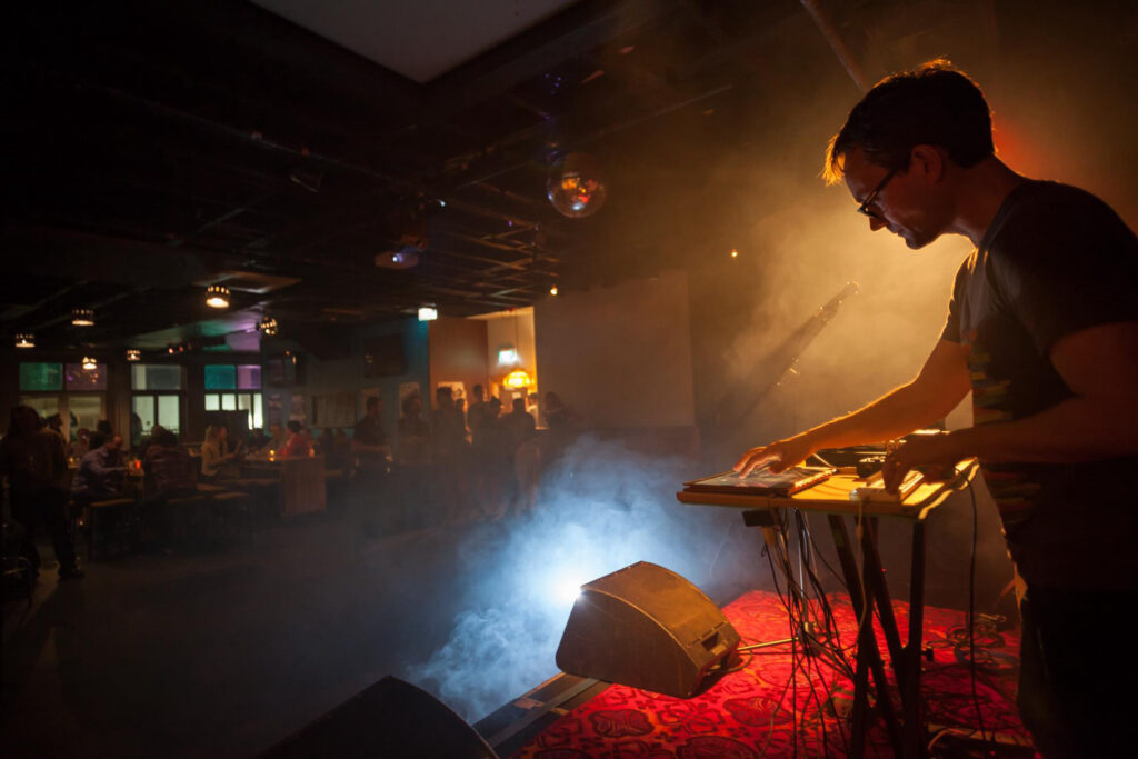 A man performing on stage with electronic music gear, in a pub.