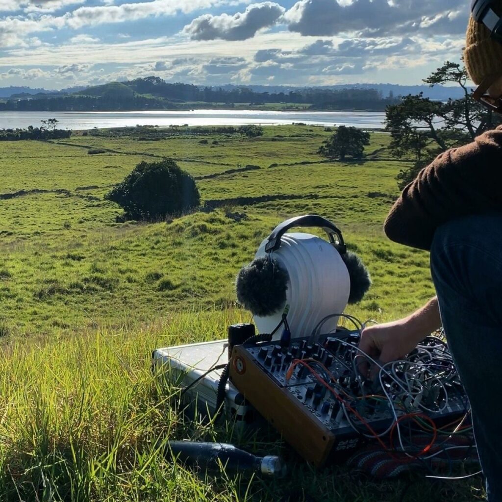 Photo of a man crouched at a box of knobs and wires on a hillside with a binaural microphone recording setup in front of him.